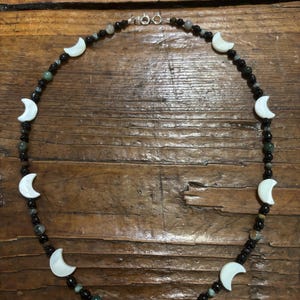 Può includere: Una collana di perline con ciondoli bianchi a forma di mezzaluna intervallati da piccole perline sferiche nere e verde scuro. La collana è disposta in forma circolare e presenta una chiusura a molla in metallo argentato nella parte superiore.