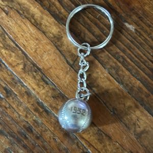 May include: A silver-toned metal keychain featuring a small, spherical baseball charm. The charm is embossed with the number 1932 and detailed with raised stitching lines. The baseball is attached to a standard circular split ring by a short chain.