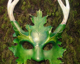 May include: A green leather mask with antlers and leaf details. The mask has a stylized face with large eyes and a pointed nose. The mask is set against a background of green moss.