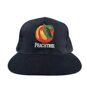May include: A black baseball cap featuring an embroidered graphic of a peach with a green leaf on the front panel. Below the peach, the word PEACHTREE is embroidered in white capital letters. The cap has a structured crown and a flat brim.