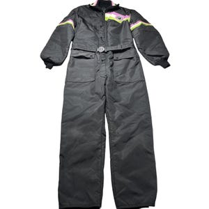 May include: A black one-piece snowsuit featuring a high collar, long sleeves with ribbed cuffs, and a belted waist. The chest area displays a chevron pattern in white, pink, and lime green, with a circular emblem centered on the upper chest.