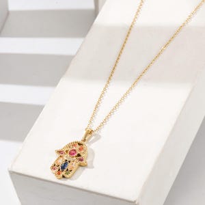 Könnte beinhalten: Goldfarbene Halskette mit einem Hamsa-Hand-Anhänger. Der Anhänger ist mit bunten Edelsteinen verziert, darunter Rot, Blau und Gelb. Die Halskette hat eine zarte Kette und wird auf einer weißen Oberfläche präsentiert.