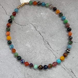 May include: A colorful beaded necklace featuring a variety of gemstone beads in shades of red, orange, yellow, green, blue, and purple. The necklace is strung on a red cord and has a gold-colored clasp. The beads are approximately 0.3 inches in diameter.