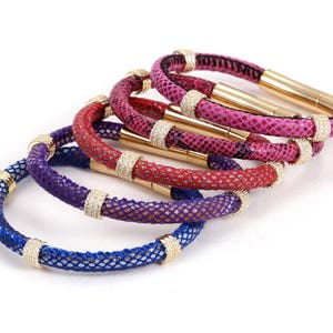 May include: A set of five colorful bracelets with a gold metal clasp. Each bracelet is made of a woven leather material with a different color: blue, purple, red, pink, and black. The bracelets are adorned with small, clear rhinestones.