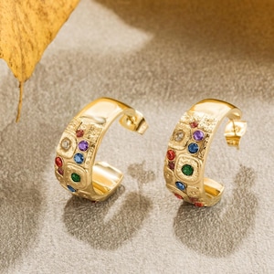 May include: A pair of gold-colored hoop earrings. Each earring features a textured surface with embedded colorful gemstones in red, blue, green, purple, and clear. The earrings have a post-back closure.