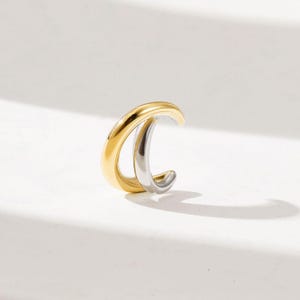 Puede incluir: Un ear cuff bicolor en oro y plata. El ear cuff tiene un diseño curvo que se envuelve alrededor de la oreja. La parte dorada está en la parte superior y la parte plateada debajo. El ear cuff es una joya moderna.