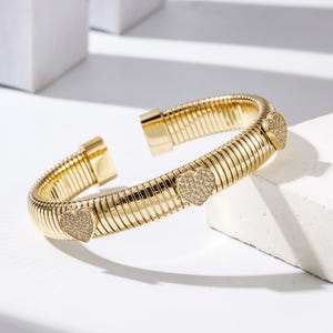 May include: A gold-colored cuff bracelet with a ribbed design. The bracelet features three heart-shaped embellishments encrusted with small, sparkling stones. The bracelet is open at one end, showcasing a clasp mechanism.