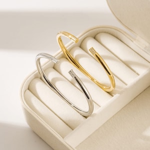 Puede incluir: Dos pulseras simples de tipo bangle con una sola fila de pequeñas piedras transparentes en cada extremo. Las pulseras se muestran en una caja blanca con forro de terciopelo blanco.