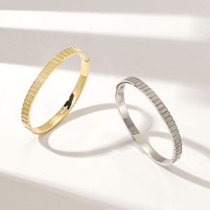 Op de afbeelding: Twee armbandjes, een gouden en een zilveren, met een vierkant ontwerp. De armbandjes zijn gemaakt van metaal en hebben een glanzende afwerking.