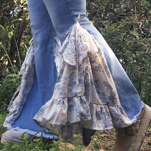 May include: Light blue denim jeans featuring flared leg openings embellished with white and blue toile de jouy patterned fabric inserts, white crochet lace trim, and fabric ties, paired with beige slip-on shoes with woven jute soles.