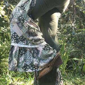 May include: Olive green trousers featuring a patchwork fabric panel on the side leg, decorated with a small floral applique at the hip and a white lace trim detail. The trousers are paired with brown wedge sandals with a textured wood-look heel.