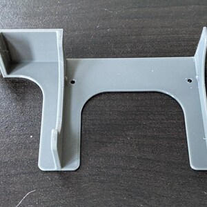 May include: A gray 3D-printed wall mount bracket featuring a rectangular frame with two vertical support arms, a side-mounted compartment, and two small circular screw holes positioned along the top horizontal bar for secure installation.