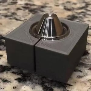 May include: A dark gray, square-shaped block composed of two interlocking pieces, featuring a centered, circular, metallic silver cone-shaped component with a smooth, polished finish and a pointed top, set into a recessed circular base.