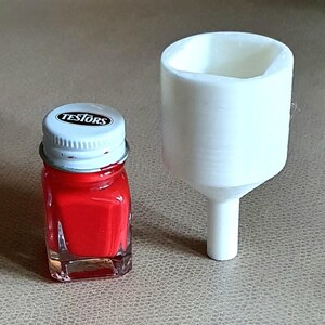 May include: A small square glass bottle of red paint with a white screw-on cap labeled TESTORS, positioned next to a white plastic funnel with a wide circular opening and a narrow cylindrical stem, both items standing upright on a flat surface.