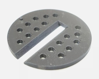 Może przedstawiać: Okrągły, czarny element wydrukowany w technologii 3D, posiadający serię małych, równomiernie rozmieszczonych okrągłych otworów ułożonych w dwa zakrzywione rzędy. Obiekt posiada wyraźne prostokątne wycięcie biegnące od środka do krawędzi.
