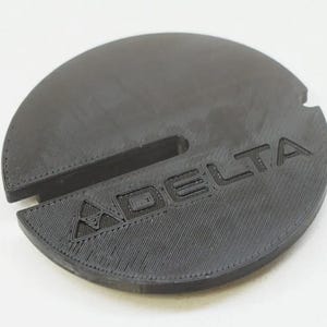 May include: A circular black plastic component featuring a textured surface with a central slot and a small notch on the edge. The word DELTA is embossed on the top surface, accompanied by a triangular logo symbol positioned before the text.