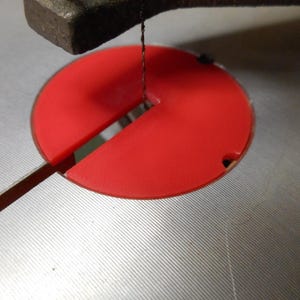 May include: A bright red circular scroll saw table insert with a central slot and a small notch on the edge, positioned within the metallic surface of a scroll saw machine, featuring a thin vertical blade passing through the center opening.