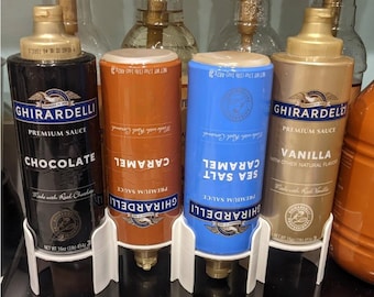 Może przedstawiać: Cztery butelki sosu premium Ghirardelli w kolorze czarnym, pomarańczowym, niebieskim i złotym, z etykietami Chocolate, Caramel, Sea Salt Caramel i Vanilla. Każda butelka jest odwrócona w białym plastikowym stojaku, z widoczną wagą netto 454g lub 482g.