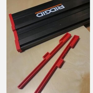 May include: A black Ridgid track saw guide rail with red end caps and two separate red plastic guide rail accessories featuring a notched design. The Ridgid brand name is printed in white text with an orange line above it on the black rail.