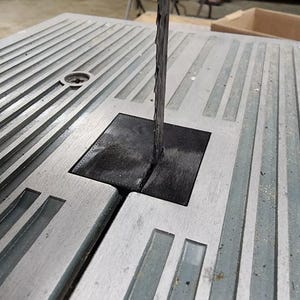 May include: A silver-colored metal bandsaw table featuring a series of parallel grooves and a central square black insert. A thin, vertical metal bandsaw blade passes through a narrow slit in the center of the black insert, extending upward from the table surface.