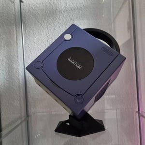 May include: A purple Nintendo GameCube video game console positioned at an angle on a black stand. The console features a circular disc cover with the Nintendo GameCube logo, a power button, a reset button, and a controller port panel on the front.