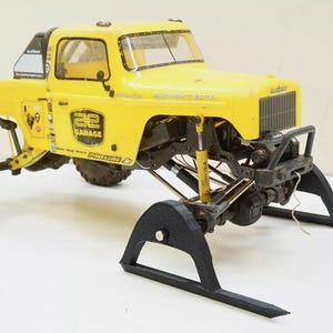May include: A yellow remote-controlled truck featuring a black roll cage, large off-road tires, and black plastic skis attached to the front axle. The body displays the text Element, Element RC, and AE Garage with various small sponsor decals.