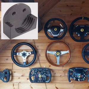 May include: A dark gray 3D-printed wall mount bracket featuring a curved, tiered interior cradle designed to securely hold a circular steering wheel. The mount has a semi-circular top section with two circular screw holes for mounting to a flat surface.