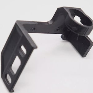 May include: A black 3D printed bracket featuring an L-shaped frame with a circular mounting ring at one end and a flat rectangular base with two elongated slots for adjustable attachment, showing visible layer lines from the manufacturing process.
