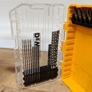 May include: A clear plastic storage case with a yellow interior, containing a set of metal jigsaw blades arranged in a black holder. The case features the partial text DEN on the front, and additional metal bits are stored in the yellow section.