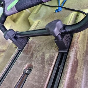 May include: A black kayak seat mounting system featuring two black plastic brackets with triangular knobs attached to a black metal frame. The brackets are secured into the molded tracks of a tan and brown textured kayak hull surface.