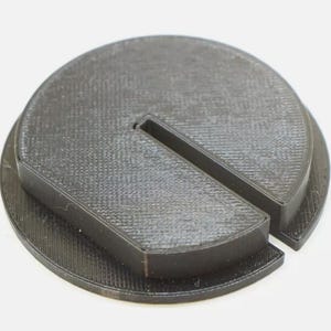 May include: A circular black object with a textured surface featuring a raised, flat-topped section that is shaped like a partial circle with a straight edge and a narrow, rectangular slot cut through the center of the raised portion.