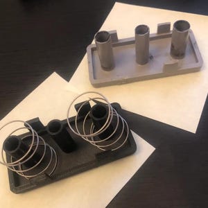 May include: Two rectangular components, one light gray and one black, each featuring three vertical cylindrical posts. The black component includes two silver metal coil springs positioned around the outer posts, while the gray component is plain.
