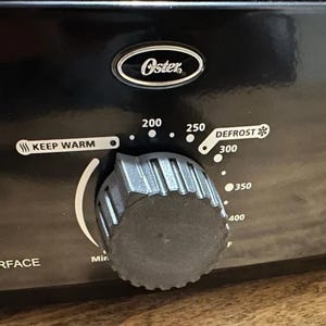 May include: A black Oster appliance control panel featuring a circular ridged knob for temperature adjustment. The panel displays settings for Keep Warm, Defrost, and numerical temperature increments from 200 to 400 degrees Fahrenheit, with a power indicator light.