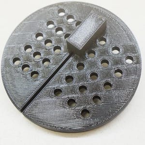 May include: A circular, black, 3D-printed disc featuring a grid of small, evenly spaced circular perforations across its surface. A rectangular block is attached to the top, and a straight, narrow slit divides the disc into two distinct halves.