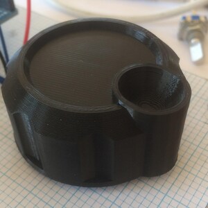 May include: A black, 3D-printed cylindrical knob featuring a large flat circular top surface and a smaller, recessed circular opening on the side. The exterior wall of the knob has vertical grooves and a layered texture from the printing process.