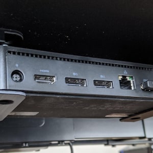 May include: A black mini computer mounted under a desk surface using a black plastic bracket. The device features a power input, an HDMI port, two DisplayPort connectors, an Ethernet port, and two USB ports with black protective covers inserted.
