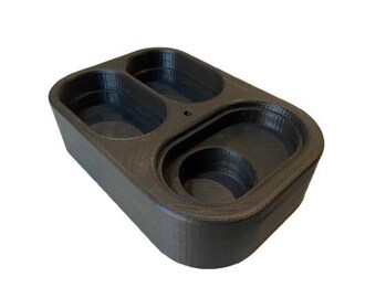 Può includere: Un vassoio organizer rettangolare nero dotato di tre scomparti incassati distinti di varie forme. Il vassoio presenta una texture liscia e stratificata tipica della stampa 3D, con angoli arrotondati e un piccolo foro circolare situato al centro.