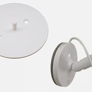 May include: A white circular wall mounting plate featuring a central peg and two screw holes, shown alongside a white wall-mounted lamp fixture with an adjustable arm and a power cord attached to a circular base with a grey edge.