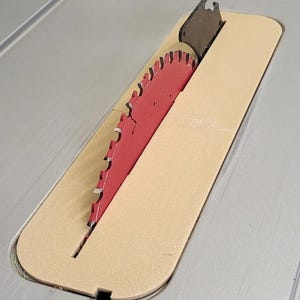 May include: A red circular saw blade with sharp teeth protruding through a beige rectangular throat plate insert. The blade is mounted on a gray table saw surface, featuring a black metal riving knife positioned directly behind the saw blade.