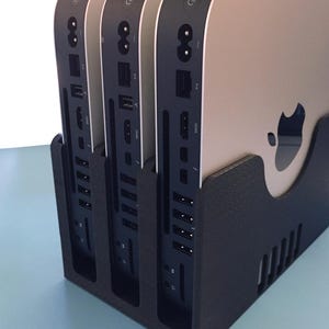 May include: Three silver Apple Mac Mini computers are arranged vertically in a black, textured, 3D-printed plastic stand. Each computer displays a black rear panel with a power button, power port, Ethernet, HDMI, and multiple USB-C and USB-A ports.