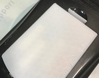 Może przedstawiać: Prostokątna biała plastikowa pokrywa z widoczną diagonalną żebrowaną teksturą, umieszczona w czarnej plastikowej obudowie. Pokrywa posiada mały zaczep na górnej krawędzi i jest osadzona równo w otaczającej ją ciemnej ramie urządzenia.