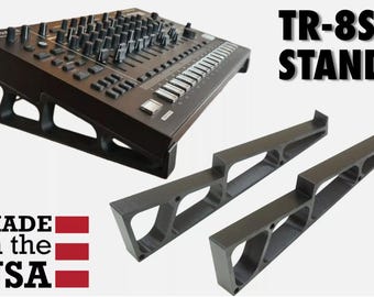 Może przedstawiać: Czarny, nachylony stojak do automatu perkusyjnego Roland TR-8S o geometrycznym wzorze wycięć. Zestaw zawiera dwie oddzielne podpórki boczne. Widoczny jest napis TR-8S STAND oraz MADE in the USA z grafiką czerwono-niebieskiej flagi.