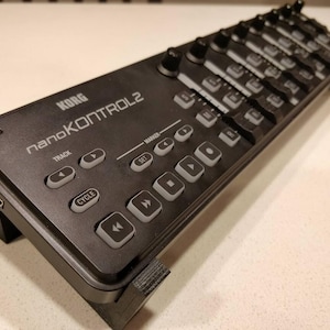 May include: A black Korg nanoKONTROL2 MIDI controller featuring a series of sliders, rotary knobs, and transport buttons. The device is mounted on a black stand and displays the brand name and model text clearly on the top surface.