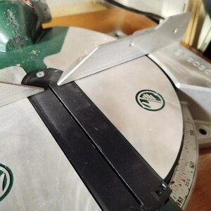 May include: A green and silver Bosch miter saw featuring a circular metal table with a central black insert. The table includes an integrated angle scale with numerical markings and a metal fence positioned across the top surface of the saw base.