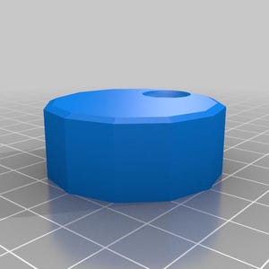 May include: A bright blue, low-profile cylindrical object with a faceted, multi-sided perimeter. The top surface is flat and features a single, circular recessed hole positioned off-center near the edge of the blue geometric form.