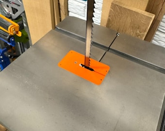 Może przedstawiać: Piła taśmowa z metalowym szarym stołem roboczym i jaskrawopomarańczową wkładką. Ząbkowane metalowe ostrze przechodzi pionowo przez środek płytki. Nad ostrzem widoczny jest niebiesko-zielony i czerwony mechaniczny zespół prowadzący.