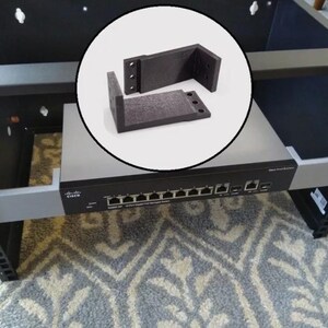 May include: A black Cisco network switch mounted in a black metal rack, featuring a circular inset showing two black L-shaped mounting brackets with three screw holes each, designed for securing networking equipment within a standard server rack.