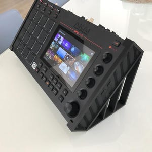 May include: A black Akai Professional MPC Live music production sampler resting on a black angled stand. The device features a central touchscreen display, a grid of sixteen square pads, multiple control knobs, and various function buttons.