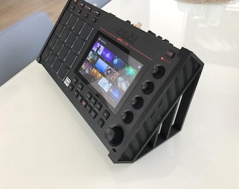 Może przedstawiać: Czarny sampler do produkcji muzycznej Akai Professional MPC Live umieszczony na czarnym, nachylonym stojaku. Urządzenie posiada centralny ekran dotykowy, siatkę szesnastu kwadratowych padów, liczne pokrętła oraz przyciski funkcyjne.
