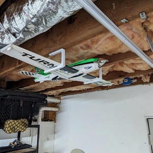 May include: A white, green, and black remote-controlled airplane model suspended from a ceiling joist by a white plastic mount. The wing features the text TURNIGY, and the fuselage displays the text AERODYNAMICS in black lettering.